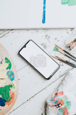 Photo for Mock up image of a mobile phone over a white table with a painting palette and painting brushes. Black mobile screen with copy space. Advertising image - Royalty Free Image