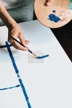 Téléchargez les photos : Gros plan de la main d'artiste âgée féminine, de la brosse à peinture et de la peinture à dessin avec peinture bleue. Peinture à l'huile colorée et émotionnelle. Peintre contemporain Création d'une œuvre abstraite moderne d'art - en image libre de droit