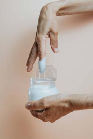 Téléchargez les photos : Old hands taking body lotion cream from a bottle, minimalist shot, pastel colour background - en image libre de droit