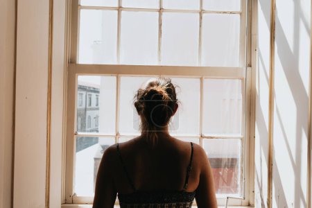 Photo for "Young woman using a dress looking through a window, giving the back to the camera, stress and anxiety concepts, dark image, sadness" - Royalty Free Image
