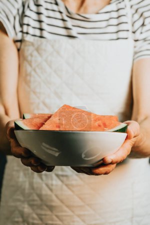 Téléchargez les photos : Old waitress offers and holds a watermelon in a dish, fruits, healthy life, good eating, mediterranean concepts, copy space, vertical image, summer eating, and refreshing fruits - en image libre de droit