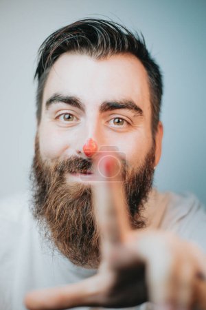 Foto de Joven hipster hombre con barba y pintura roja en la nariz sonriendo y jugando retrato, concepto de artista joven, primer plano retrato, imagen divertida, creatividad y diversión - Imagen libre de derechos