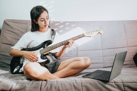 Foto de Image of happy beautiful woman playing guitar and composing song. Copy space. African woman learning new skills. Watching the lesson on the laptop. - Imagen libre de derechos