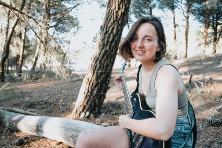 Photo for Young hipster woman playing the guitar smiling outside the forest park of the city. Having fun learning a new skill, music play seasonal style. Young short hair girl. Copy space - Royalty Free Image