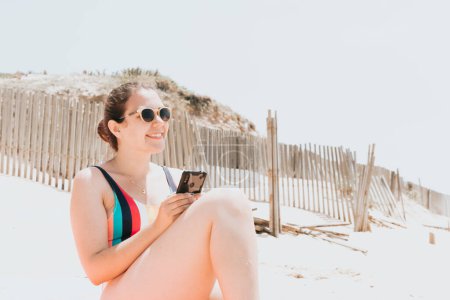 Téléchargez les photos : "Jeune femme sourit tout en bavardant au téléphone portant un maillot de bain coloré à la plage prenant un bain de soleil, voyage concept jeune vacances, copier l'espace, réseau social, concept d'utilisateur de lunettes de soleil" - en image libre de droit