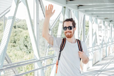 Téléchargez les photos : Portrait of a young man saluting someone at the airport or bus station , luggage, bags and suitcase. Smiling hipster traveler with sunglasses, copy space, sunny day - en image libre de droit