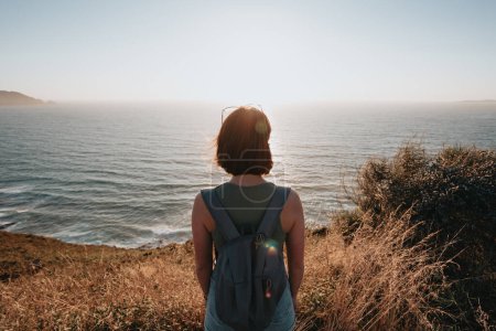 Photo for Back image of a young backpack traveler hiker during an stunning sunset coastline scenario, hipster traveler concept, beauty landscape. Copy space for text. Movement and liberty, nomad life. - Royalty Free Image