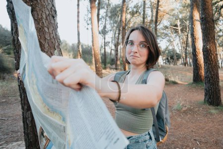Foto de "Young woman traveler with backpack walking among trees at forest with a map traveling. concept of travel. cannot find right way, wears optical spectacles, dressed in casual clothes" - Imagen libre de derechos