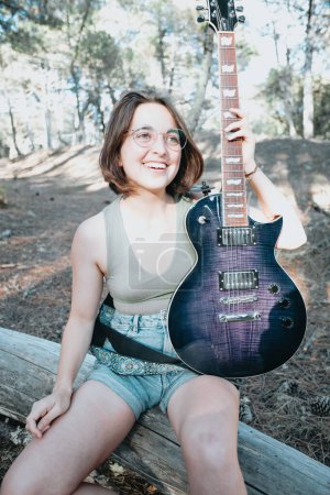 Téléchargez les photos : Portrait of an smiling young hipster woman holding and playing the guitar outside the forest park of the city. Having fun learning a new skill, music play seasonal style. Copy space - en image libre de droit