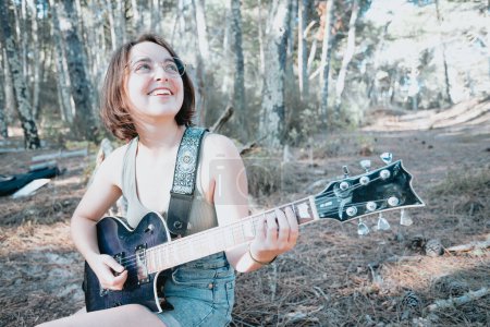 Photo for Young hipster woman sitting in a trunk playing guitar on park or garden background. Teen girl learning to play song and writing music. Hobby, lifestyle, relax, Instrument, leisure, education concept - Royalty Free Image