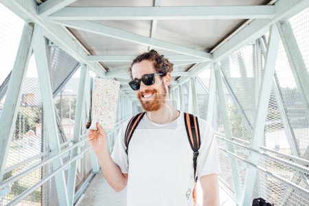 Photo for "Portrait of a young man at the airport or bus station , luggage, bags and suitcase holding map. Smiling hipster traveler with sunglasses, copy space, sunny day" - Royalty Free Image