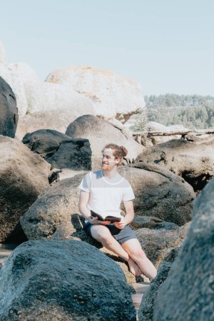 Photo for Young man looking away from camera while reading a book on the shore of the sea, copy space, confidence and study concepts, future and self help - Royalty Free Image
