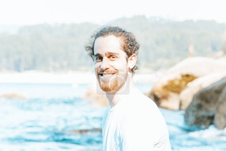 Téléchargez les photos : Jeune hipster mâle aux longs cheveux bouclés et à la barbe souriant à la caméra lors d'une journée ensoleillée à la plage, le bonheur et les concepts de détente - en image libre de droit