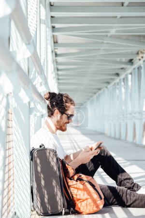 Foto de "Young hipster man at the airport or bus station waiting while writes a text message with the luggage, bags and suitcase. Young man long hair hipster traveler with sunglasses, copy space, sunny day" - Imagen libre de derechos