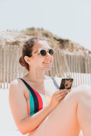Téléchargez les photos : "Young woman smiles while chatting on the phone wearing a colorful swimsuit at the beach taking a sun bath, travel young holiday concept, copy space, social network, sunglasses user concept, close up" - en image libre de droit