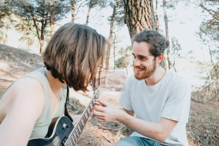 Foto de Handsome Young Man Teaching hipster Girl to Play Guitar on the forest trees during a sunset. Learning a new skill joyful concept. Music with copy space. Young couple electric guitar - Imagen libre de derechos