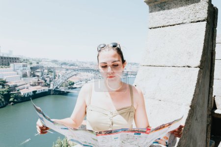 Téléchargez les photos : "Young woman tourist using sunglasses looking a map in the middle of a street and smiling thinking to camera, summer day, sunny, porto, mediterranean city," - en image libre de droit