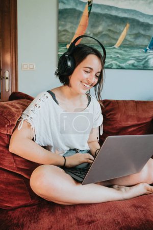 Foto de "Mujer joven feliz en un paño relajado trabaja en casa en un sofá rojo con un ordenador portátil y escuchar música, trabajo remoto y la educación, quedarse en casa durante un virus. Sonriente vida feliz en el interior." - Imagen libre de derechos