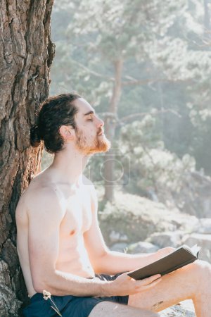 Téléchargez les photos : Close up of a man with a beard holding a book and relaxing shirtless next to a tree during summertime, vertical image - en image libre de droit