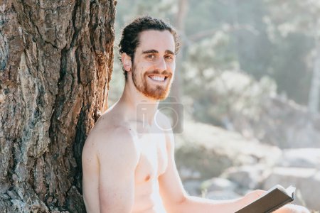 Foto de Close up of a man with a beard holding a book and smiling to camera shirtless next to a tree during summertime - Imagen libre de derechos