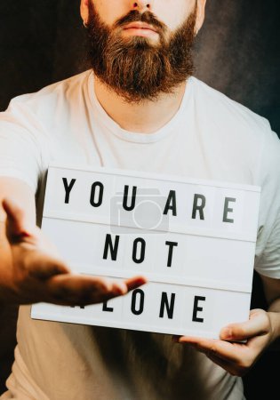 Photo for Young bearded hipster man holding a sign that says you are not alone, giving hands to the camera, help and self help concept, mental health - Royalty Free Image