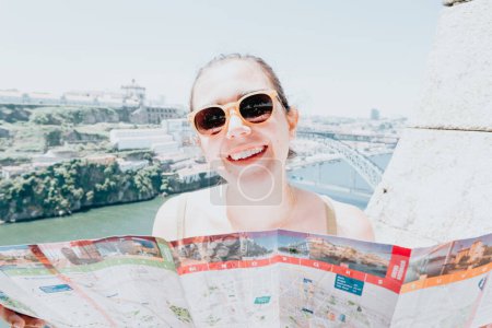 Photo for "Young woman tourist using sunglasses looking a map in the middle of a street and smiling thinking to camera, summer day, sunny, porto, mediterranean city," - Royalty Free Image