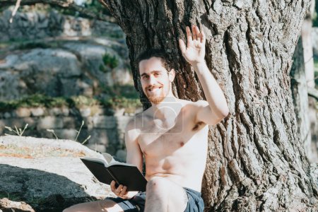 Téléchargez les photos : "Un jeune hippie en malles torse nu lisant un livre contre un arbre et saluant à la caméra pendant une journée d'été, se détendre et se détendre concepts, bonne vie, les jeunes" - en image libre de droit