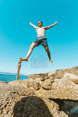 Téléchargez les photos : Jeune homme aux cheveux longs sautant dans les airs, torse nu lors d'une journée ensoleillée, concept d'espace et de liberté, vacances, image fantastique, journée à la plage - en image libre de droit