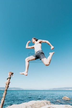 Foto de Joven macho con el pelo largo saltando en la playa, sin camisa durante un día soleado, concepto de espacio y libertad, vacaciones, imagen fantástica, corriendo en el aire - Imagen libre de derechos