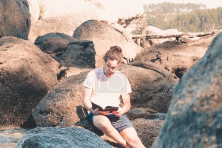 Téléchargez les photos : Jeune homme hippie sur une chemise blanche lisant un livre dans le rocher de la plage pendant une journée d'été, vacances froid et liberté - en image libre de droit