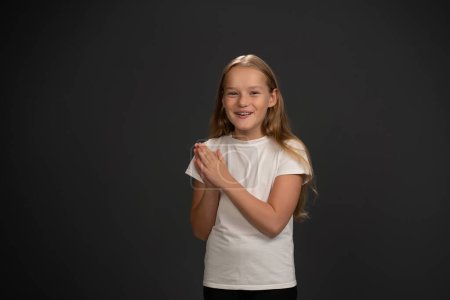 Téléchargez les photos : Petite fille excitée inspirée par les parents avec les mains réunies regardant joyeusement la caméra portant un t-shirt blanc et un pantalon noir isolé sur fond noir - en image libre de droit