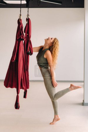 Téléchargez les photos : Femme heureuse fait du yoga sur un hamac bourgogne suspendu dans une salle de gym lumineuse - en image libre de droit