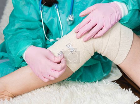 Foto de Un médico que usa guantes médicos envuelve una venda elástica alrededor de la pierna y la rodilla del paciente. El traumatólogo aplica un material elástico al paciente - Imagen libre de derechos