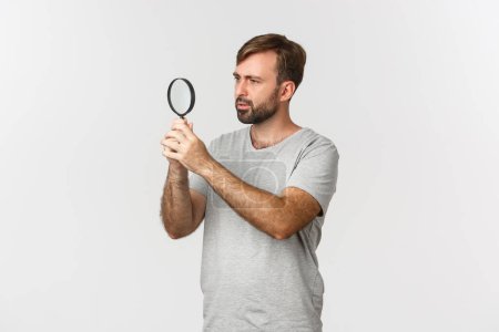 Foto de Imagen de hombre caucásico guapo en camiseta gris, buscando algo, mirando a través de lupa con cara curiosa, de pie sobre fondo blanco - Imagen libre de derechos