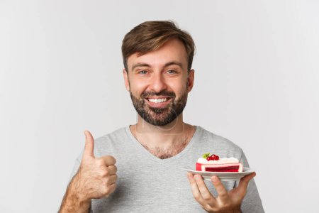 Téléchargez les photos : Gros plan d'un homme caucasien joyeux avec barbe, portant un t-shirt gris, tenant un gâteau et montrant les pouces, comme un dessert, debout sur fond blanc - en image libre de droit