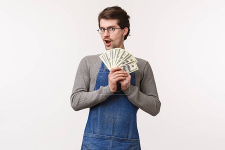 Photo for Small business, finance and career concept. Enthusiastic young male student working part-time at coffee shop, wear apron holding his salary, fan of dollars, winning money, white background - Royalty Free Image