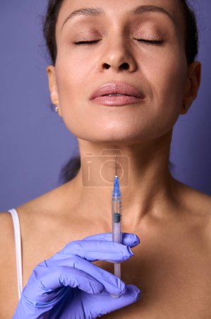 Photo for Close-up beauty portrait of middle aged pretty woman posing on purple background with a syringe of cosmetic rejuvenating product in her hand. Injection cosmetology, anti-aging treatment concept - Royalty Free Image