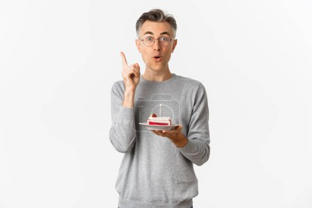 Photo for Image of happy middle-aged man, celebrating birthday, have great idea what to wish while blowing candle on b-day cake, standing over white background - Royalty Free Image