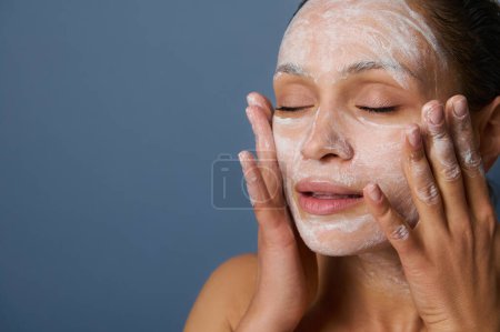 Téléchargez les photos : Beautiful woman massaging her face while removing make-up using a foam cleansing cosmetic product, and refreshing her skin with an exfoliant beauty product, isolated over gray background. Close-up. - en image libre de droit