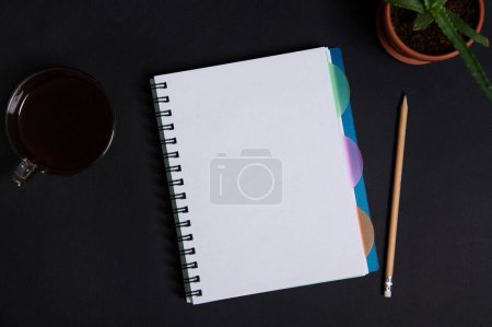 Photo for A potted succulent in clay pot, cup of coffee and wooden pencil lying down next to a copybook, notepad, organizer with white blank sheet with space for text, on black table background. - Royalty Free Image