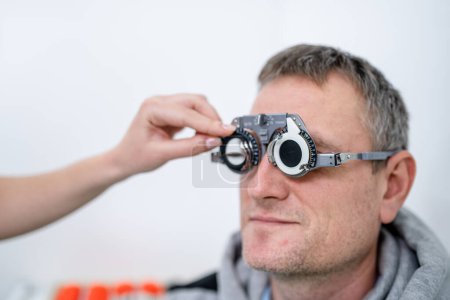 Photo for "Male patient undergoes an eye test and prescription for eyeglasses in ophthalmology clinic. Optometrist checking patient eyesight and vision correction. Changing lenses on trial frame on patient nose" - Royalty Free Image