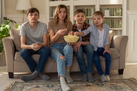 Photo for Happy family, a woman and three boys, are sitting on the sofa in the living room with a bowl of popcorn, watching TV - Royalty Free Image