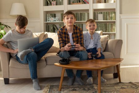 Photo for Three brother boys playing computer games on a laptop and video games with joysticks in their hands - Royalty Free Image