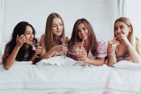 Foto de Beber algunos buenos cócteles. Chicas alegres en la ropa de dormir acostado en la cama en la habitación blanca y tener celebración - Imagen libre de derechos