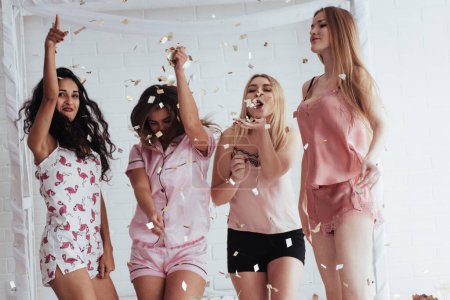 Photo for "Party is going good. Confetti in the air. Young girls have fun on the white bed in nice room" - Royalty Free Image