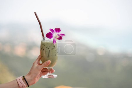 Photo for "Hand holding green tea matcha latte cold beverage drink with orchid flower." - Royalty Free Image