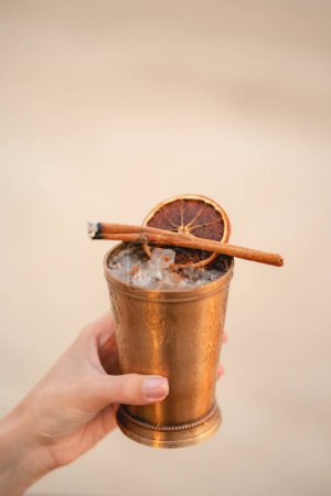 Foto de Mano con cóctel, Bebida alcohólica con hielo, naranja y canela en la playa. - Imagen libre de derechos