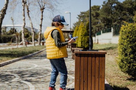 Téléchargez les photos : "Schoolboy kid throwing the trash into dumpster. Boy using recycling bin to throw away the litter. Caucasian child recycles the junk into the trash-can" - en image libre de droit