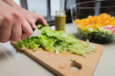 Photo for "Close-up of a girl's hand cut a salad on a wooden table, a woman prepares a vegetarian salad, healthy food, a knife cuts greens" - Royalty Free Image