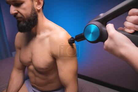 Photo for "Bearded caucasian man getting percussion shoulder massage by a massager in a professional massage parlour. shock wave muscle recovery technique" - Royalty Free Image